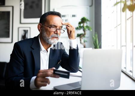 Serious confused mature european man in suit works at laptop, uses credit card, has problems with investing Stock Photo