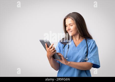 Happy senior european woman doctor in blue uniform typing on digital tablet, posing on light gray background, free space Stock Photo