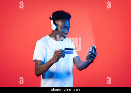 Amazed young black guy using phone, headphones and bank card Stock Photo