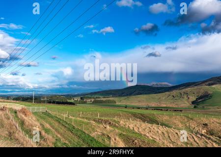 Photograph of a large rainbow over a green agricultural field with cows and sheep grazing and a mountain range in the background in New Zealand Stock Photo