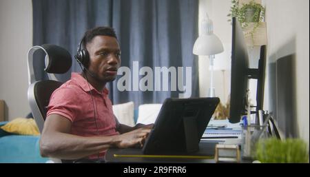 African American web designer or developer works remotely at home. Man sits and talks on online conference at computer. Tablet and multiple monitors stands on table. Concept of working at cozy home. Stock Photo