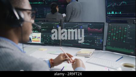 Team of financial analysts analyze exchange market charts on multiple big screens. Shot changing on African American man working at computer with displayed real-time stocks. Trading and investment. Stock Photo