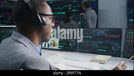 Team of financial analysts analyze exchange market charts on multiple big screens. Shot changing on African American man working at computer with displayed real-time stocks. Trading and investment. Stock Photo