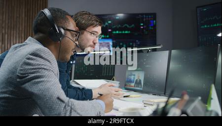 Multiracial financial analysts talk and monitor real-time stocks on computer. Businessmen work in bank office. Big digital screen with exchange market charts. Cryptocurrency trading and analytics. Stock Photo