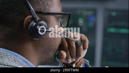 African American financial analyst in headset working in bank office. Businessman looks at computer screen, monitors real-time stocks, exchange market charts. Investment and analytics. Close up. Stock Photo