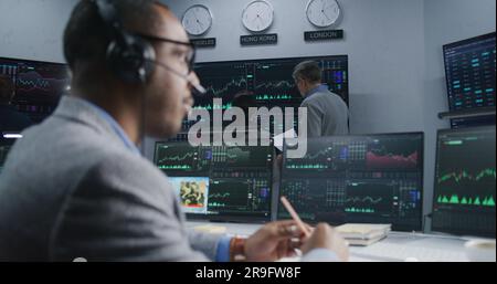 Team of financial analysts analyze exchange market charts on multiple big screens. Shot changing on African American man working at computer with displayed real-time stocks. Trading and investment. Stock Photo