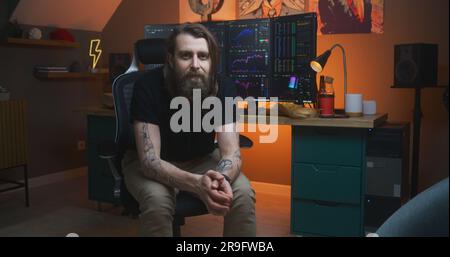Male stock trader, freelancer sits and looks at camera. Multi-monitor computer workstation with real-time stocks, exchange market charts. Man works in investment at home office. Cryptocurrency trading Stock Photo