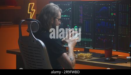 Trader surfs internet using phone while taking break from analyzing real-time stocks, exchange market charts on multi-monitor computer. Man works in investment at home office. Cryptocurrency trading. Stock Photo