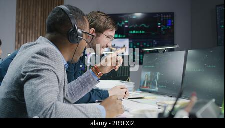 Multiracial financial analysts talk and monitor real-time stocks on computer. Businessmen work in bank office. Big digital screen with exchange market charts. Cryptocurrency trading and analytics. Stock Photo