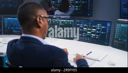 Software programmer sits in front of computer with blockchain network and data server in monitoring office. Team of IT technical specialists work with live analysis charts on big digital screens. Stock Photo