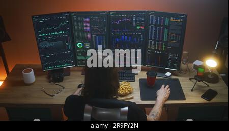 Back view of male stock trader watching real-time stocks, exchange market charts on multi-monitor computer workstation, drinking coffee. Man works in investment at home office. Cryptocurrency trading. Stock Photo