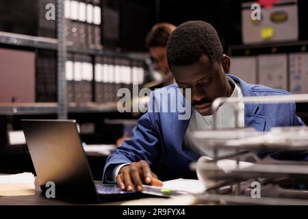 Businessman analyzing administrative files, searching for bureaucracy record in corporate depository. African american bookkeeper working late at night at accountancy report Stock Photo