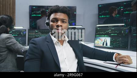 African American financial analyst works at computer with displayed real-time stocks, turns and looks at camera. Colleagues analyze exchange market charts at background. Investment concept. Portrait. Stock Photo