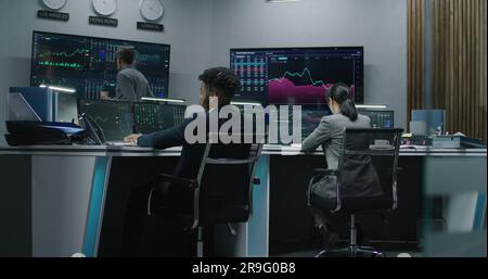 Multi ethnic team of traders work on multi-monitor computers with displayed real-time stocks. Colleagues analyze exchange market charts on multiple big screens. Cryptocurrency trading and investment. Stock Photo