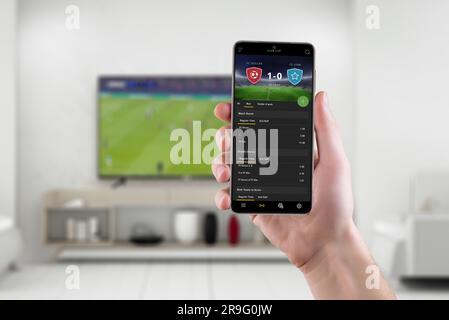 Live soccer betting experience in the comfort of a living room with smart phone. Phone with modern betting app interface in hand. Soccer match broadca Stock Photo