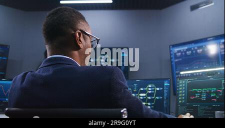 Software programmer sits in front of computer with blockchain network and data server in monitoring office. Team of IT technical specialists work with live analysis charts on big digital screens. Stock Photo