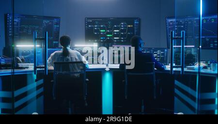 Back view of diverse IT technical specialists working on computers with database server in modern monitoring office at night. Big digital screens with real-time analysis charts and blockchain network. Stock Photo