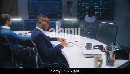 Software programmer sits in front of computer with blockchain network database in monitoring control room. Team of IT technical specialists work with real-time analysis charts on big digital screens. Stock Photo