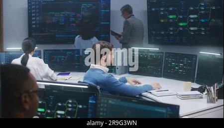 Technical support specialists sit at computers with displayed blockchain network map and server data. Big data scientists work in monitoring room. Big screens on the wall. Concept of cyber security. Stock Photo