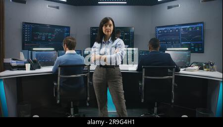 Female big data scientist stands in monitoring room and looks at camera. Software engineers work at computers at background. Multiple big screens with displayed real-time analysis charts. Dolly shot. Stock Photo