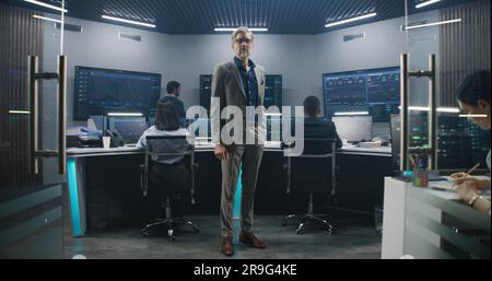 Mature big data scientist puts on glasses, stands in monitoring room and looks at camera. Software engineers work at computers at background. Multiple big screens with displayed live analysis feed. Stock Photo