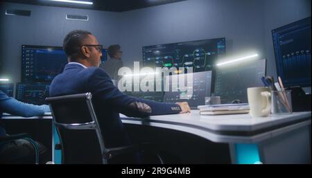 Multiethnic IT technical specialists sit in front of computers with data server in monitoring office. Software engineer works with live analysis charts and blockchain network on big digital screens. Stock Photo