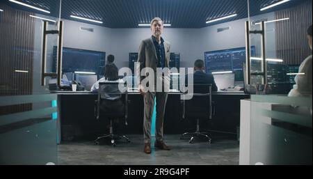 Mature big data scientist puts on glasses, stands in monitoring room and looks at camera. Software engineers work at computers at background. Multiple big screens with displayed live analysis feed. Stock Photo
