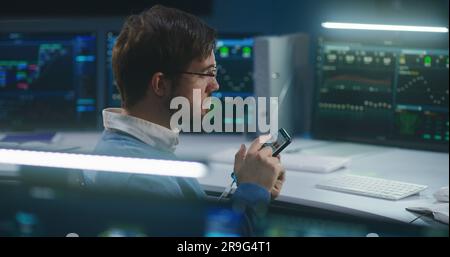 Professional IT technical specialist chats using phone, sits in front of computer with big data server, blockchain network and live analysis charts, works in monitoring office. Cyber security concept. Stock Photo