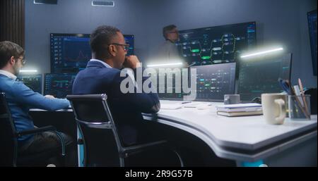 Multiethnic IT technical specialists sit in front of computers with data server in monitoring office. Software engineer works with live analysis charts and blockchain network on big digital screens. Stock Photo