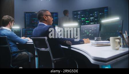 Multiethnic IT technical specialists sit in front of computers with data server in monitoring office. Software engineer works with live analysis charts and blockchain network on big digital screens. Stock Photo