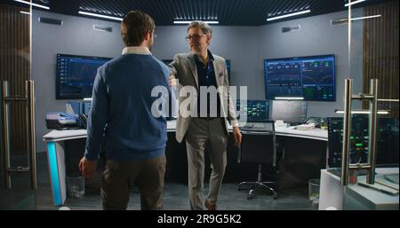 Mature supervisor in business suit greets programmers in monitoring room and looks at camera. Big data engineers start work. Multiple big screens with displayed real-time analysis charts. Dolly shot. Stock Photo
