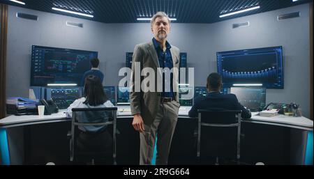 Mature big data scientist puts on glasses, stands in monitoring room and looks at camera. Software engineers work at computers at background. Multiple big screens with displayed live analysis feed. Stock Photo