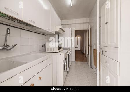 Old kitchen with mismatched white cabinets, white square tiles, loose white appliances, white tile sink, white wooden door with opaque yellow glass Stock Photo