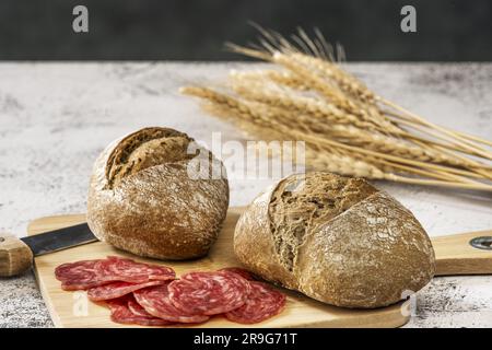 Still life with village bread on a wooden board together with a knife and slices of salami sausage on a white surface Stock Photo