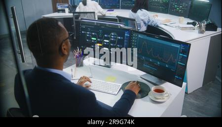 African American big data scientist works at computer in monitoring room. Multiracial colleagues analyze real-time charts at background. Multiple big screens on the wall with displayed server data. Stock Photo