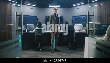 Mature big data scientist puts on glasses, stands in monitoring room and looks at camera. Software engineers work at computers at background. Multiple big screens with displayed live analysis feed. Stock Photo