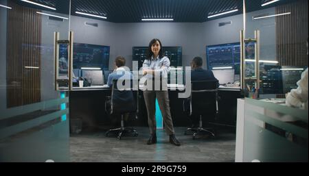 Female big data scientist stands in monitoring room and looks at camera. Software engineers work at computers at background. Multiple big screens with displayed real-time analysis charts. Dolly shot. Stock Photo