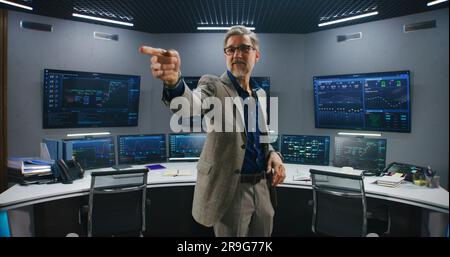 Mature supervisor in business suit greets programmers in monitoring room and looks at camera. Big data engineers start work. Multiple big screens with displayed real-time analysis charts. Dolly shot. Stock Photo