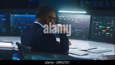 African American IT technical specialist works on computer with data server and real-time analysis charts in modern monitoring office. Team of software engineers and big digital screens on background. Stock Photo