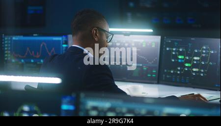 African American software engineer sits in front of computer and big digital screens with data server and blockchain network, works in modern monitoring control room with real-time analysis charts. Stock Photo