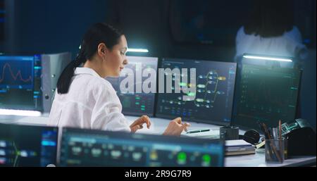 Female IT technical specialist works on computer with data server and blockchain network database in modern monitoring control room. Team of software engineers and big digital screens on background. Stock Photo