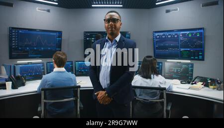 African American IT scientist stands in monitoring room and looks at camera. Multi cultural big data engineers work at computers. Multiple screens with displayed real-time analysis charts. Dolly shot. Stock Photo