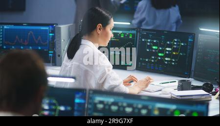 Female IT technical specialist works on computer with data server and blockchain network database in modern monitoring control room. Team of software engineers and big digital screens on background. Stock Photo