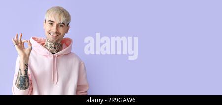 Young tattooed man in hoodie showing OK on lilac background with space for text Stock Photo