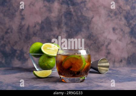 Glass of cold Cuba Libre cocktail and bowl with limes on purple background Stock Photo