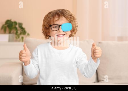 Happy boy with eye patch on glasses showing thumbs up indoors. Strabismus treatment Stock Photo