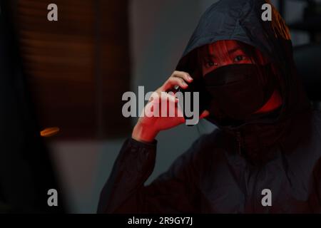 Hacker in hoodie talking on smartphone and typing code to hacking programming system or server. Stock Photo
