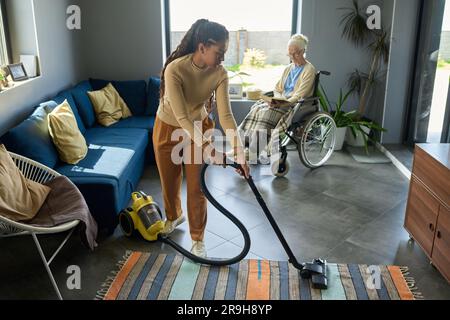 Youthful girl with vacuum cleaner helping her grandmother with disability with domestic chores while senior woman reading book by window Stock Photo