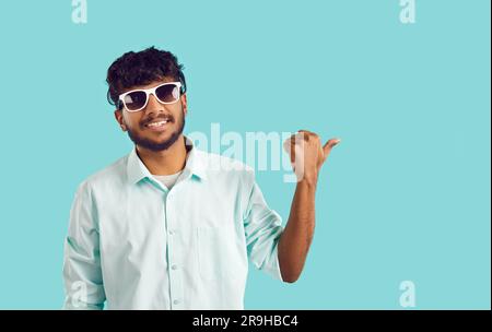 Young Indian man in sunglasses smiling pointing with thumb at empty space stands in studio Stock Photo