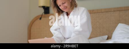 Young woman in white bathrobe working remotely with laptop in bed at home Stock Photo